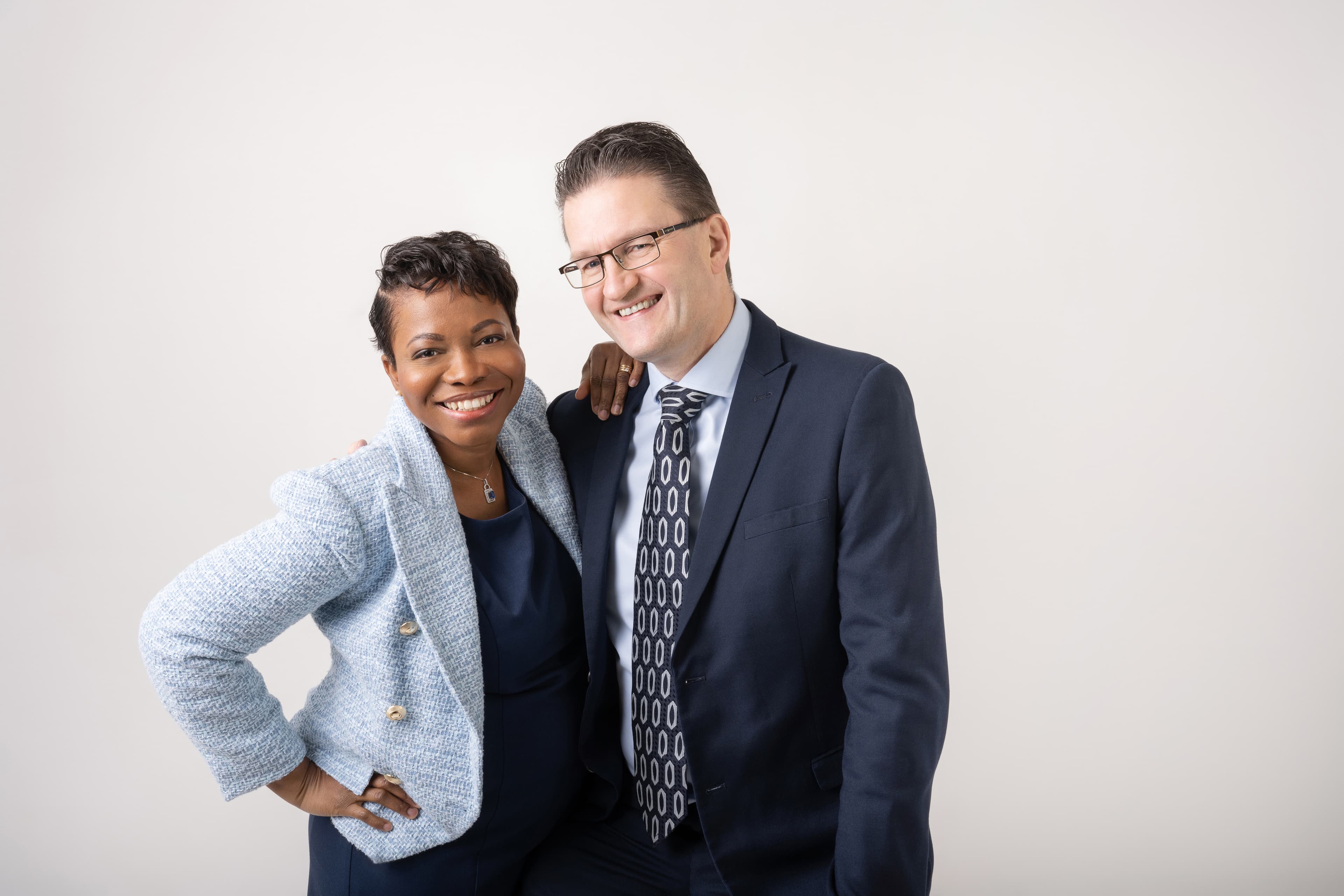 Smiling Black woman in blue tweed blazer and white man in suit posing together.