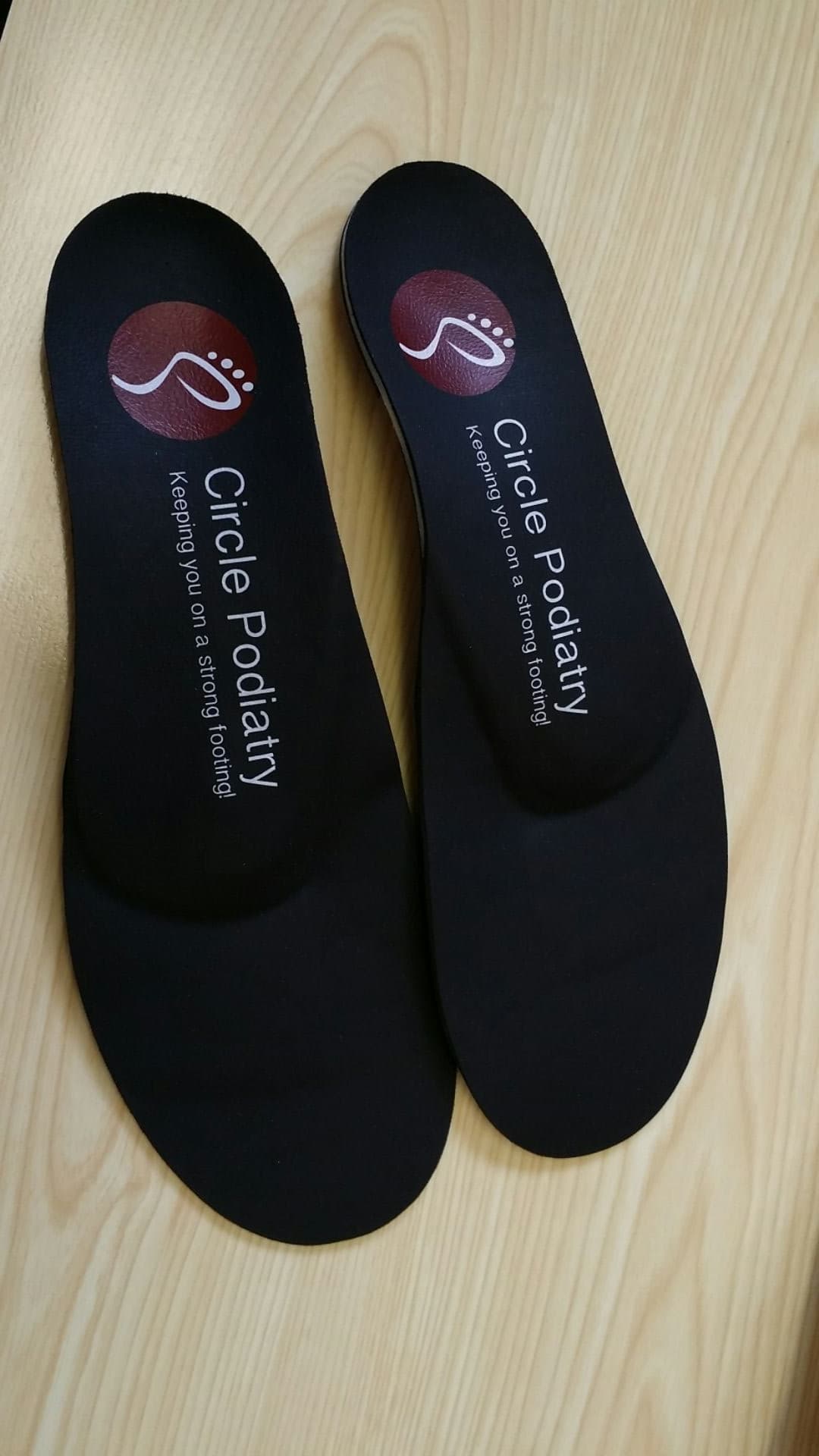 Two black orthotic insoles with a red Circle Podiatry logo on a wooden surface.