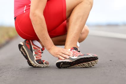 A runner in red shorts crouches on the road, clutching a painful ankle injury.
